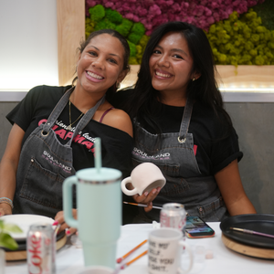 Two smiling women wearing matching gray denim aprons with 'SOULLAND LAB' printed on them, seated at a table with mugs, paintbrushes, and cans in a bright indoor setting.