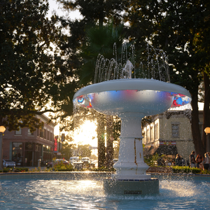 A white outdoor fountain in a town square at sunset, with water spraying upward and colorful light reflections on the basin. Trees, buildings, and a few people are visible in the background.