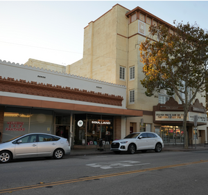 A wide street scene with storefronts along the left side, a beige multi-story building in the center, parked cars, and a tree in the foreground under a clear sky.