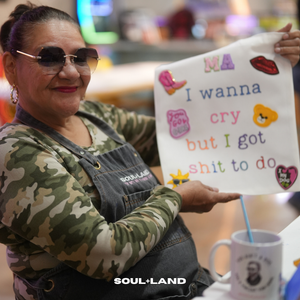 A woman wearing a camouflage shirt and denim overalls holds up a white sign with colorful text and stickers. The sign reads 'MA I wanna cry but I got shit to do' with small decorative graphics.
